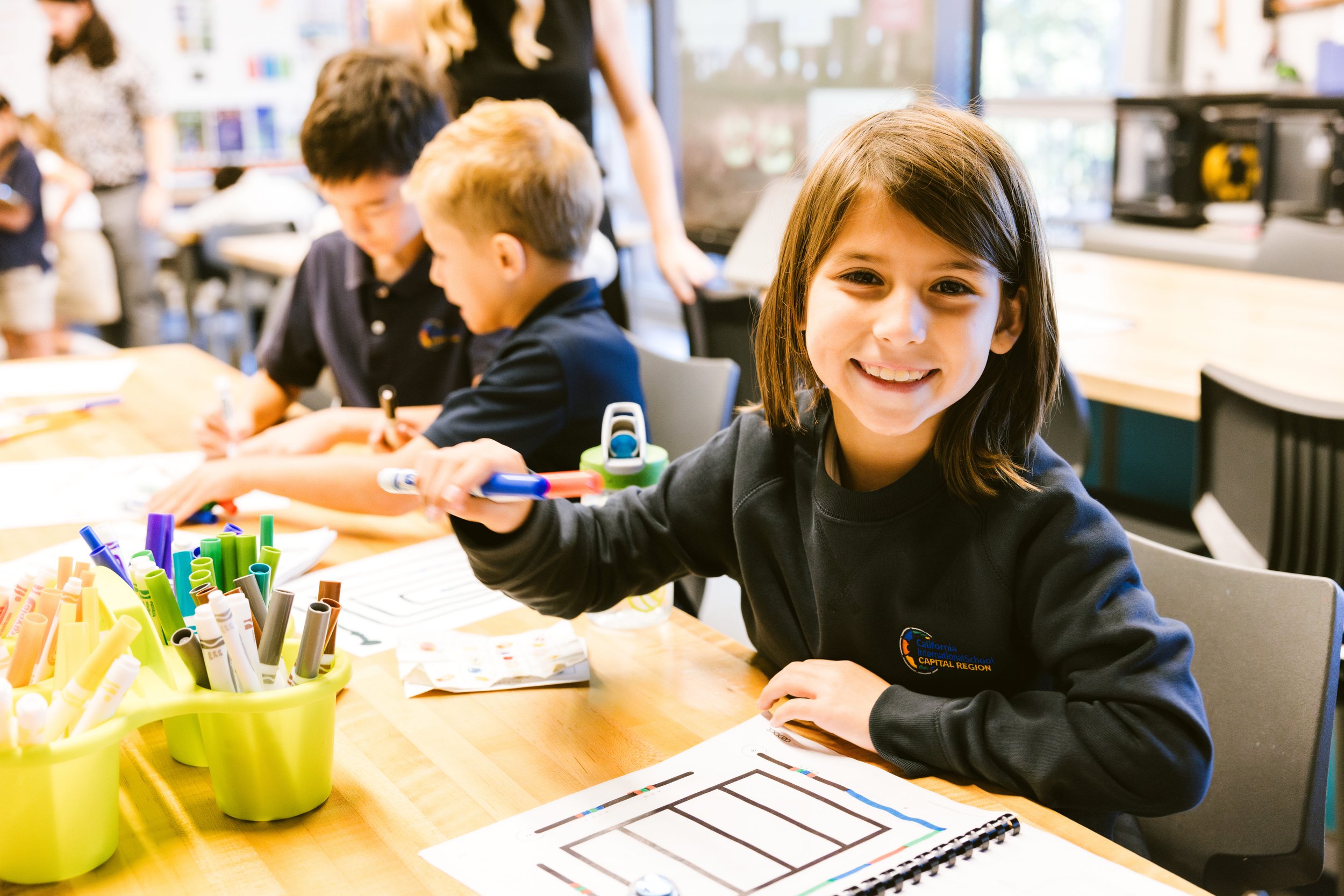 A girl smiling at her desk, holding up a pencil.