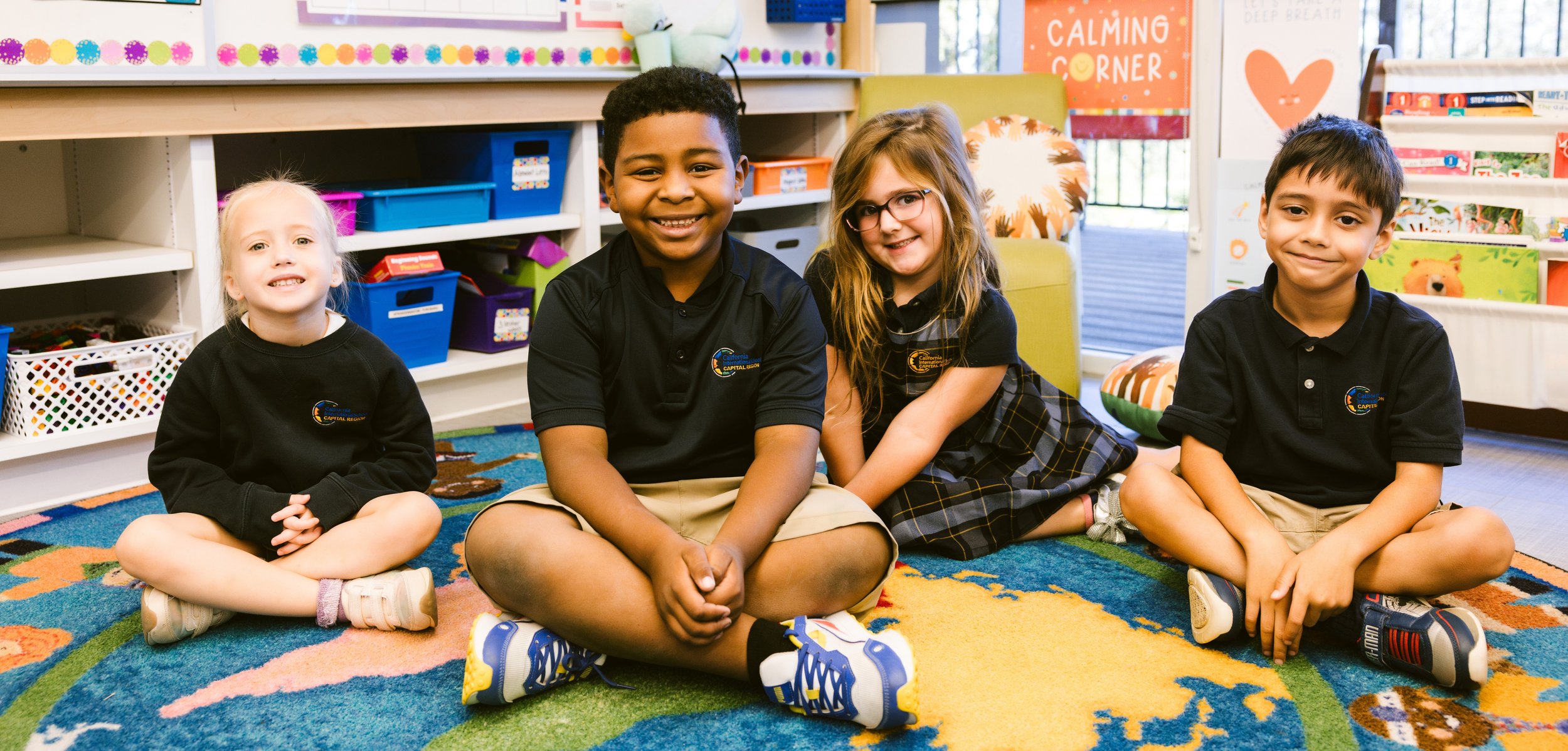 Four diverse elementary school students sitting together on a colorful classroom carpet, smiling at the camera - representing CIS's inclusive learning community