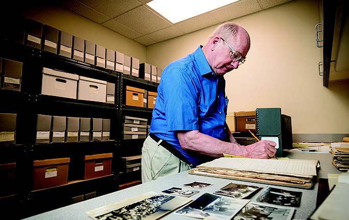 Teigen writing in an old book down in the archives room; shelves of boxes behind him