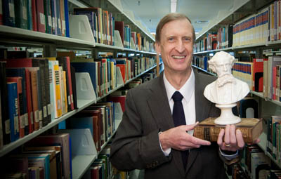 Dr. Robert Hanna poses with a plaster bust of Charles Dickens in Memorial Library among the bookshelves..