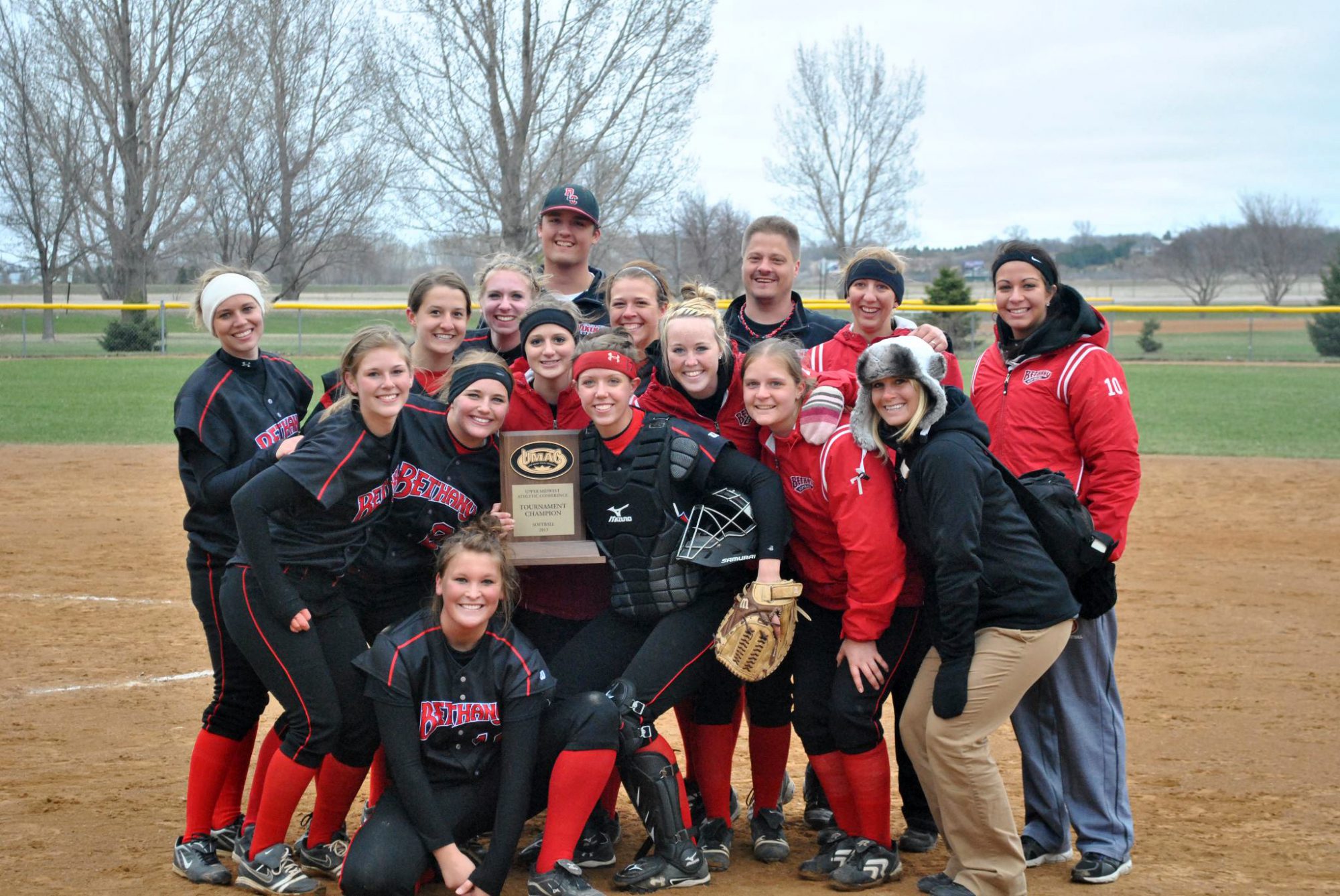 Women's softball team pose with tournament champion trophy on the field