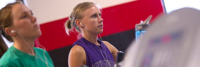 female students with earbuds in using exercise equipment in fitness center