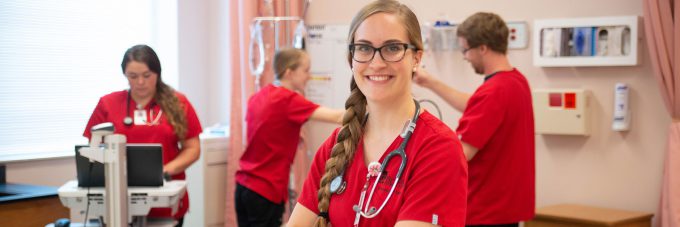 female nursing student in red scrubs and stethoscope smiling at camera; three other nursing students in red scrubs working in background