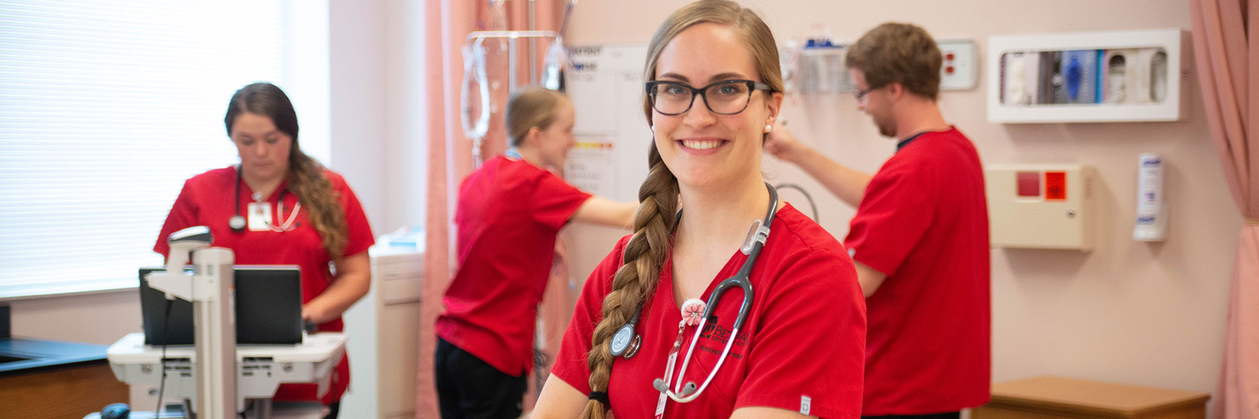 female nursing student in red scrubs and stethoscope smiling at camera; three other nursing students in red scrubs working in background