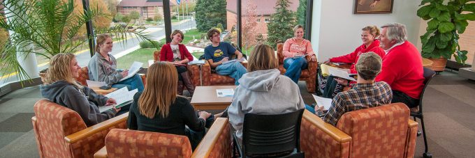 students and a professor sitting in chairs around a circle in room with large windows overlooking campus