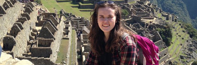 Female student sitting on rock wall with ancient spanish ruins on hillside behind her