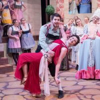 Man in german outfit poses with woman in greek goddess dress; all other women on stage watch - dressed in costumes from many different time periods.