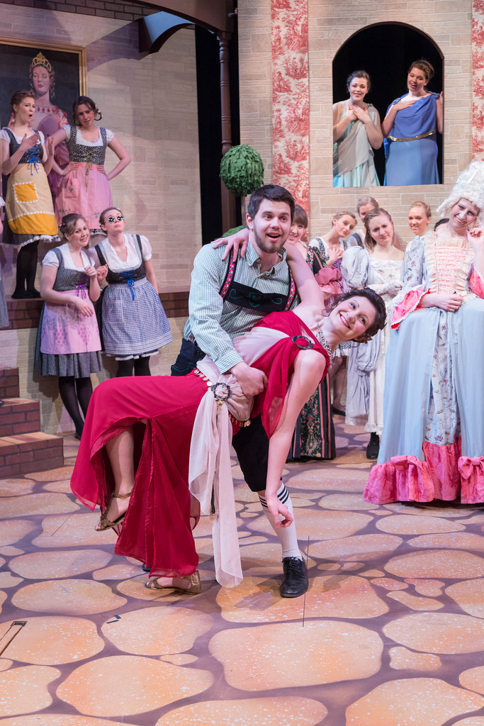 Man in german outfit poses with woman in greek goddess dress; all other women on stage watch - dressed in costumes from many different time periods.