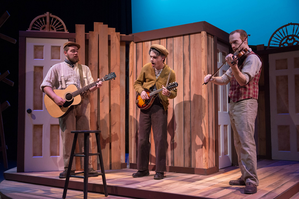 Three men playing instruments and singing on stage in front of a wooden fence