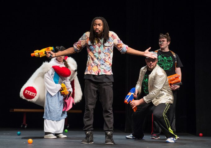 Man on stage holding a nerf gun, shrugs his shoulders; other 3 actors on stage behind him