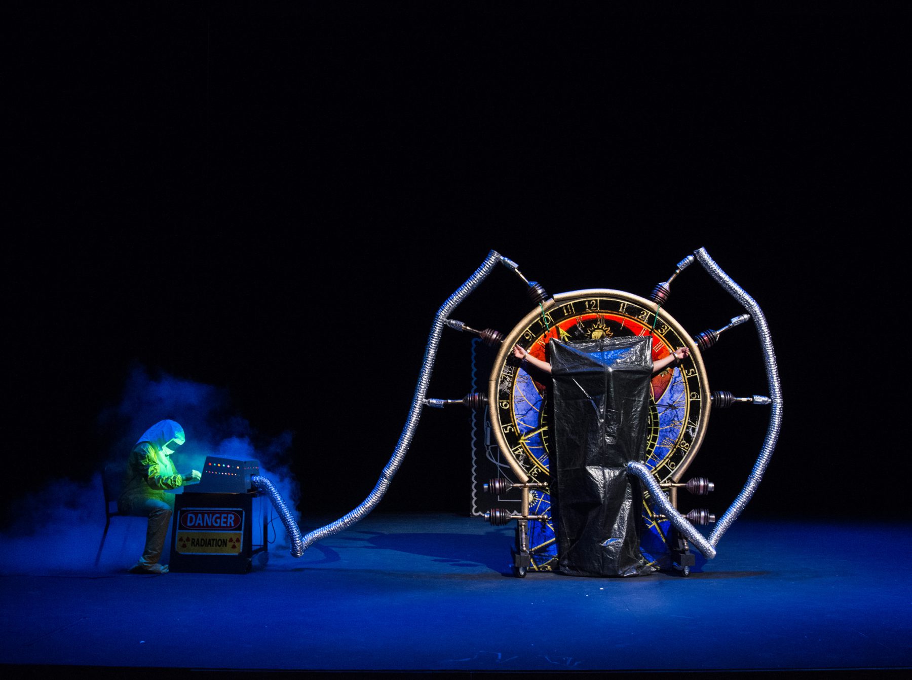 Person in Hazmat suit sits at a machine that says "Danger Radiation" and is connected to a device to which a person is attached and covered up (Theatre physics)