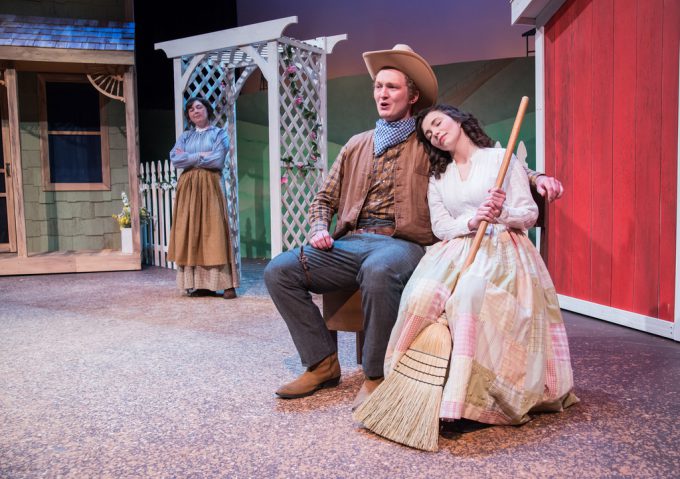 Cowboy (actor) and female actor sit on bench together while other female actor looks at them from across stage