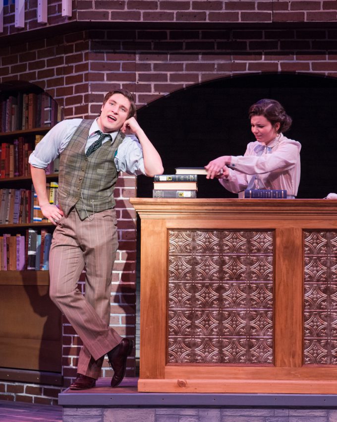 Male actor with elbow on stack of books talks while female actor behind desk moves the books