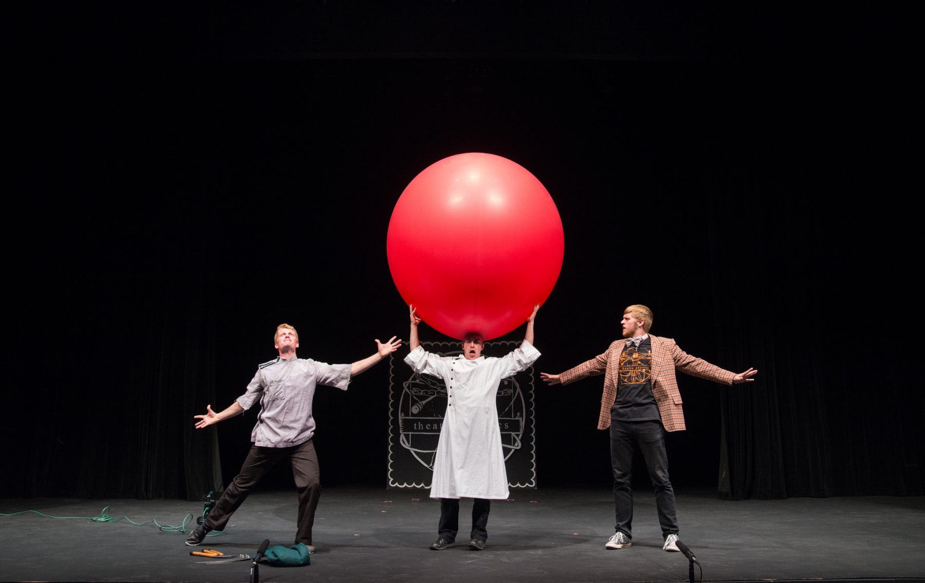 Scientist holds giant red ball on head; two men on either side of him