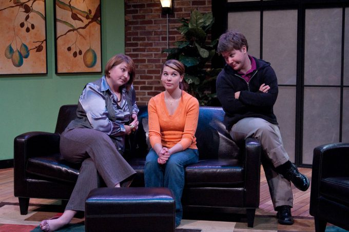 Two female actors sit on a couch; male actor sits on arm of couch. Stage made to look like a therapist's office