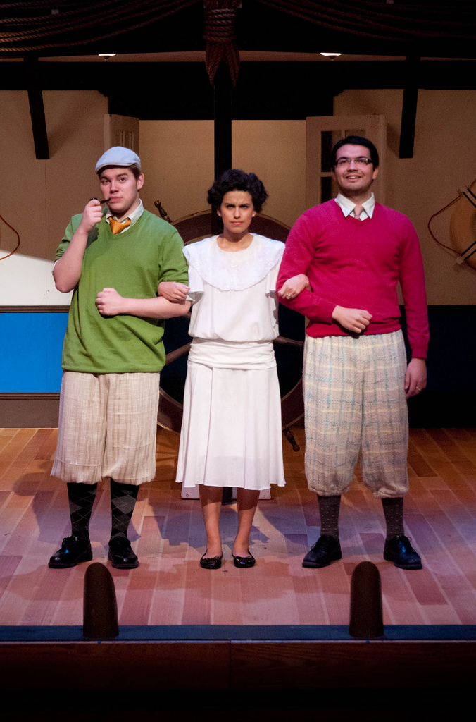 Three actors on stage. Two men dressed in knickers, shirt, tie, and sweater. Female in middle in white dress