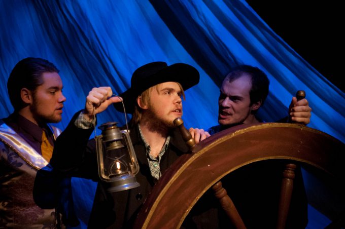 Three actors stand behind a large wooden ship wheel