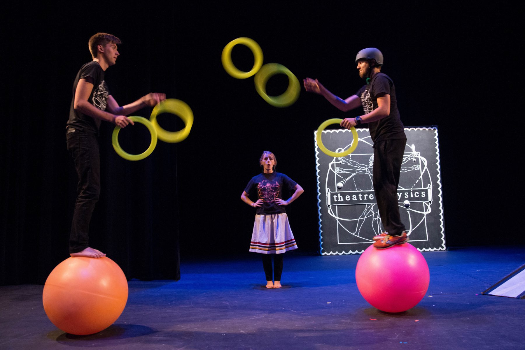 Two men standing on yoga balls and juggling yellow rings while a female student observes