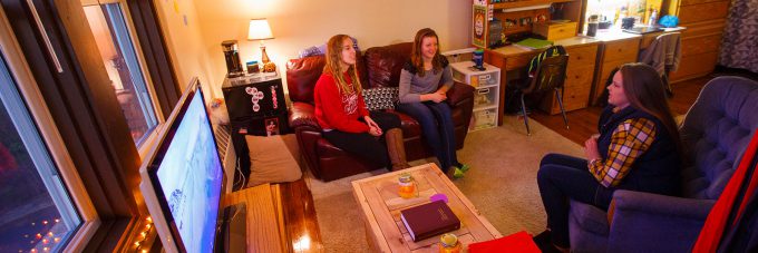 Three girls sitting in dorm room talking; 2 on couch, one in chair.