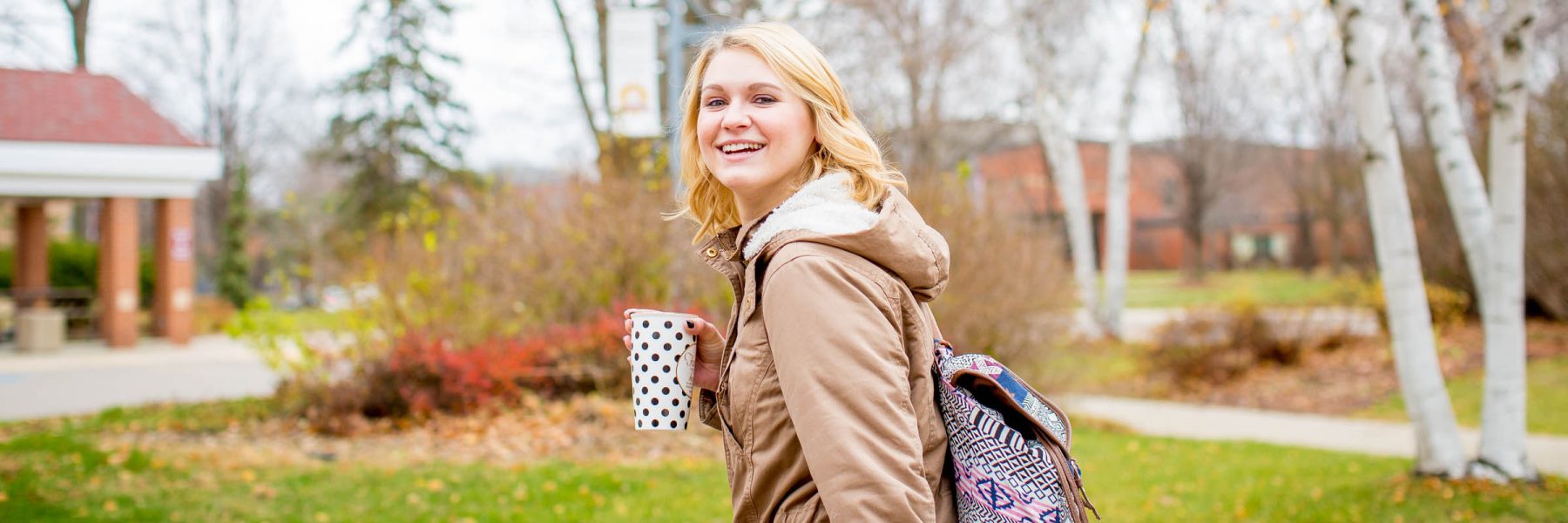 Female student wearing coat and backpack, carries a coffee mug as she walks across campus