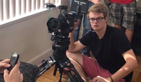 Student sits on floor behind video camera to record a shot