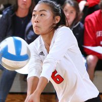 A volleyball player bumps a ball during a game in the gym. Fans on the bleachers look on.