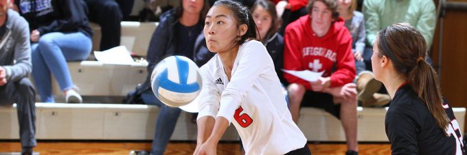 A volleyball player bumps a ball during a game in the gym. Fans on the bleachers look on.