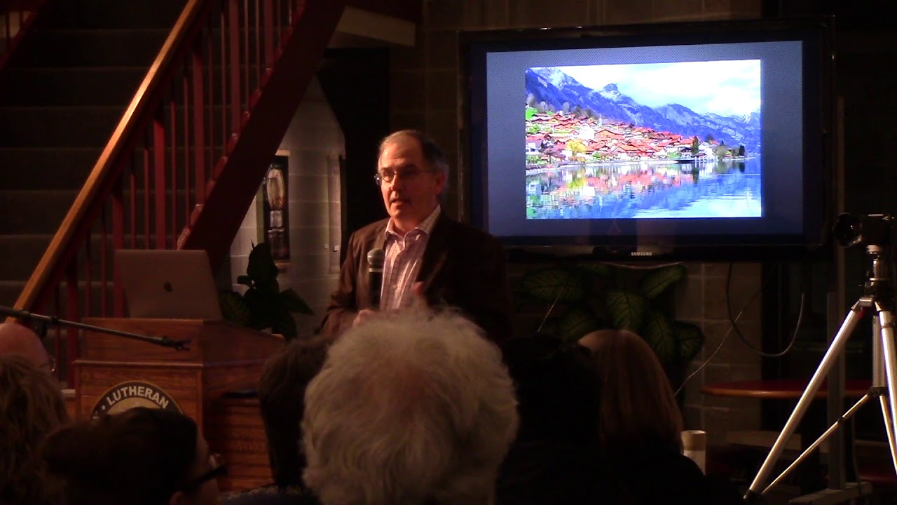 Guest artist Wes Taylor speaks at the podium during an art lecture in the Ylvisaker Fine Arts Center gallery, with his art displayed in the background.