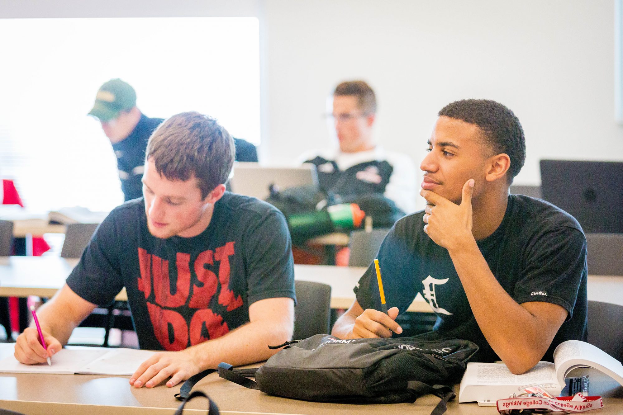 Two male students in class; one taking notes, one looking at the speaker