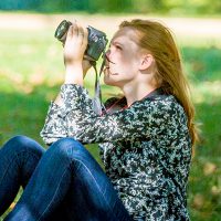 Student sitting on ground outside on a sunny day photographing into the trees.