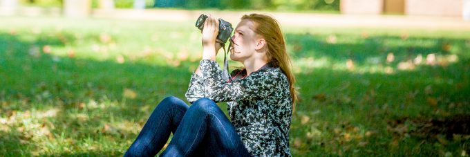 Student sitting on ground outside on a sunny day photographing into the trees.