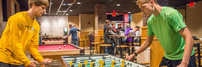 students play a game of foosball in a room with a pool table and others mingling