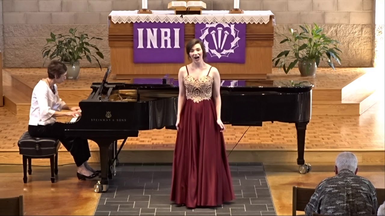 Female soloist in long red dress sings in front of the piano, played by Prof. Balge, in front of the chapel