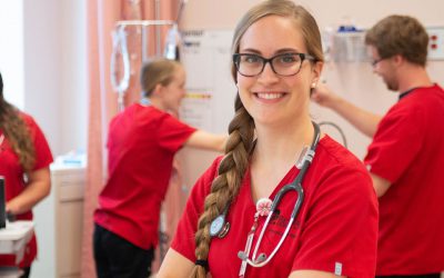 A female nursing student wearing red scrubs and a stethescope smiles at the camera while other nursing students work in the background