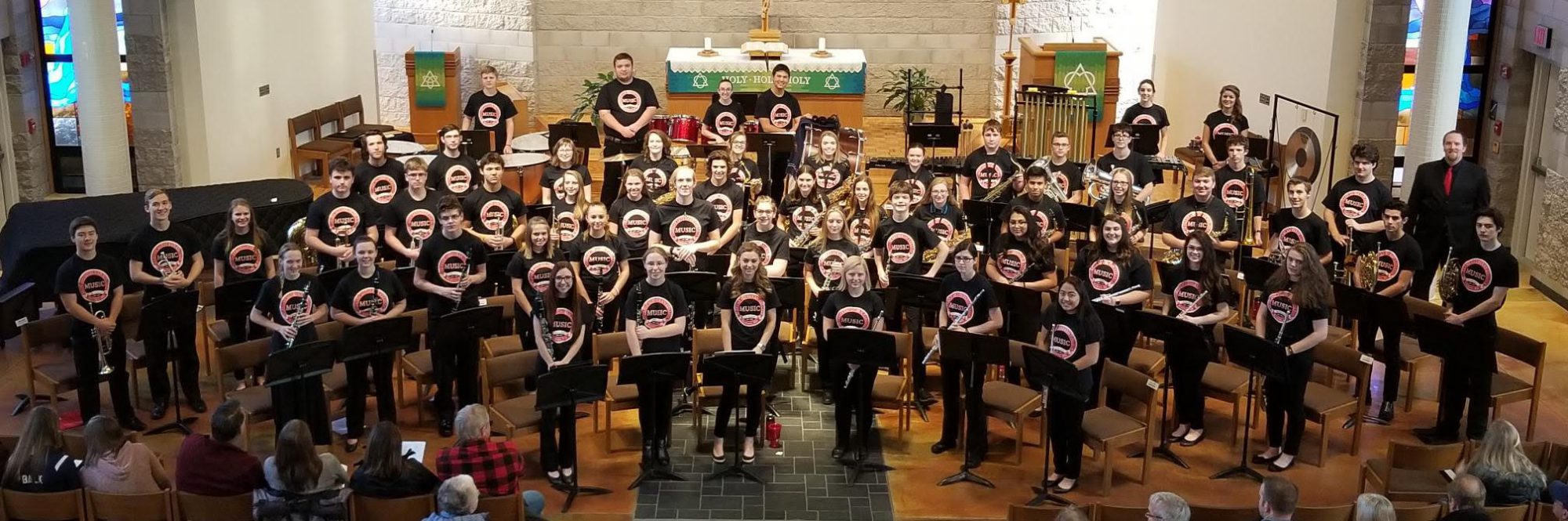 High school and college band members stand with instruments in front of chapel