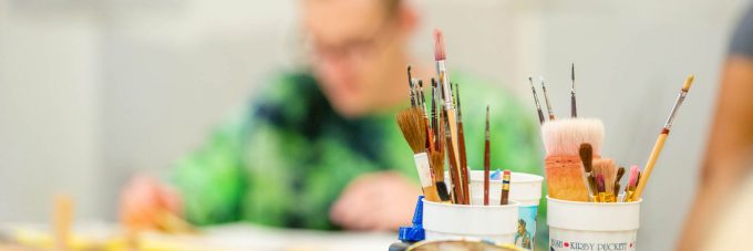 various sizes of paint brushes are sitting in cups while a blurred out student works in the background