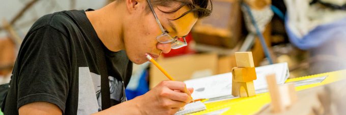 male student draws with a pencil on paper with wooden blocks on his desk