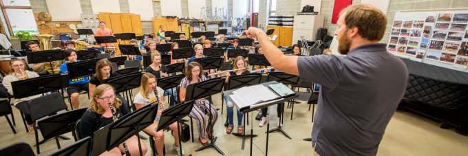 band director stands and conducts a rehearsal of students seated behind music stands while playing instruments