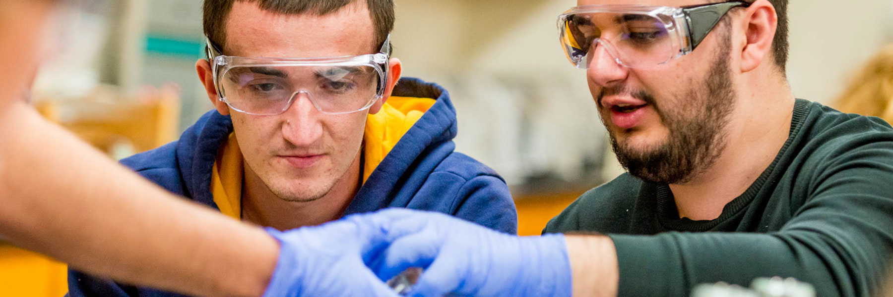 two male students with goggles and protective gloves handle objects in chemistry class