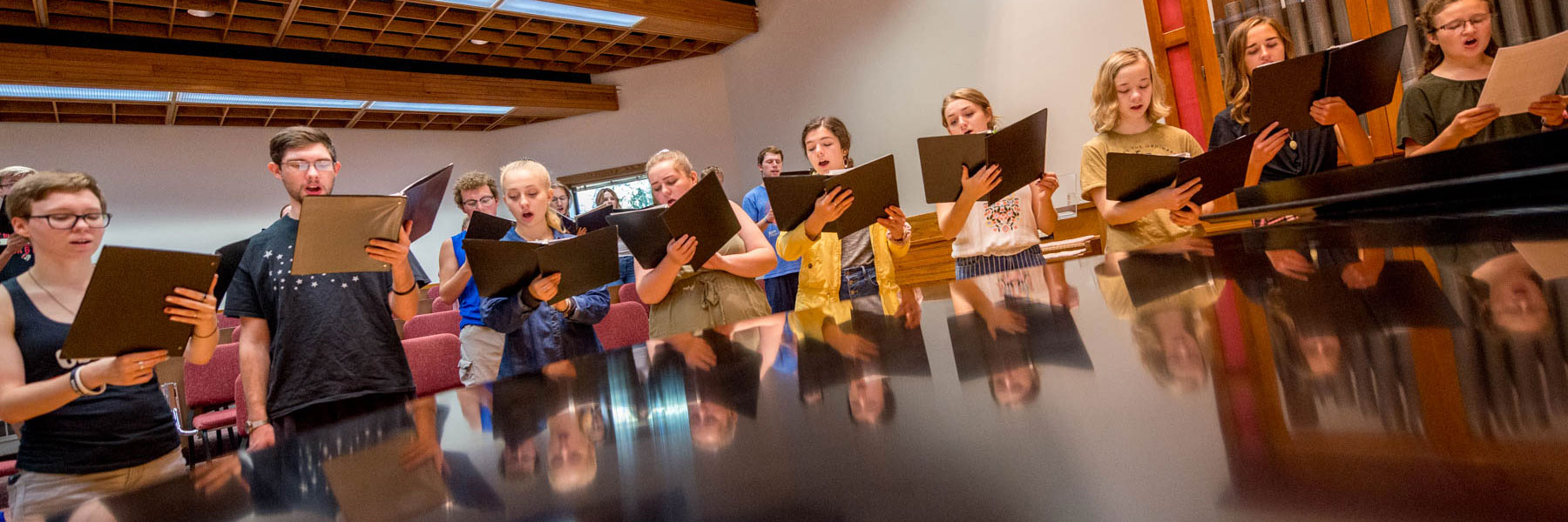 students hold folders and sing around a piano during a choir rehearsal