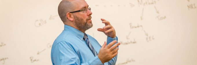 a male professor gestures with his hands as he lectures to students