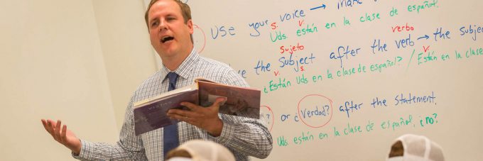 a professor talks while holding a book and gesturing with his hands during a spanish class