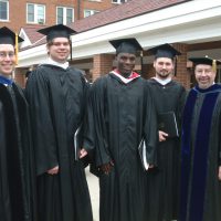 4 male graduates and a professor in graduation gowns outside of Old Main