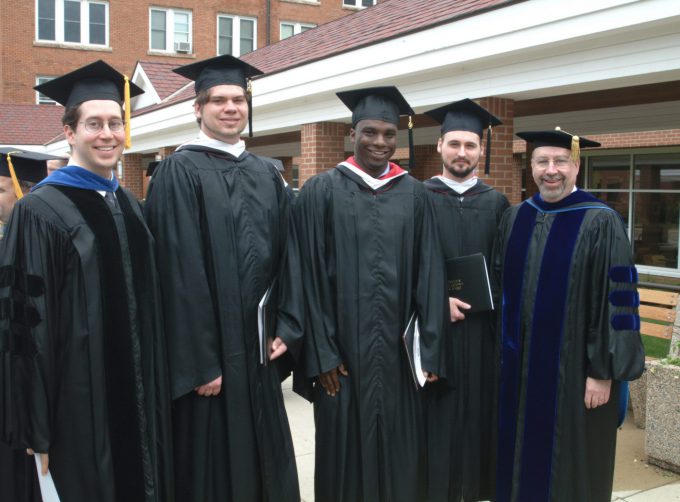 4 male graduates and a professor in graduation gowns outside of Old Main