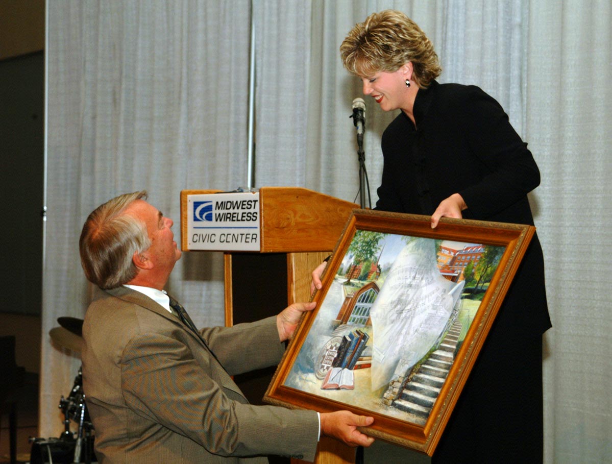 Woman on stage hands a painting to a man standing on floor in front of stage