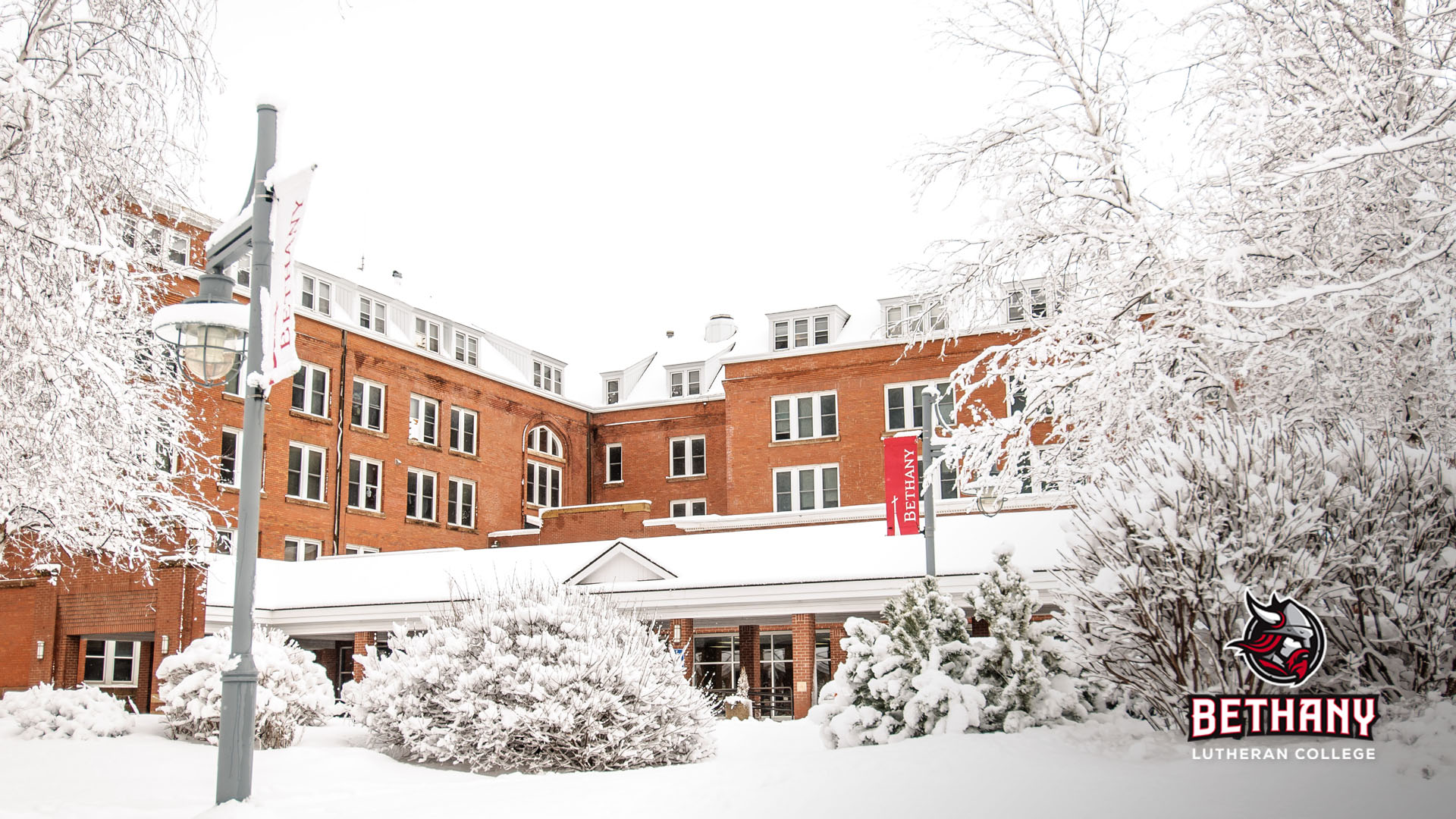 Snow-covered trees and shrubs outside of Old Main after a fresh snowfall. Bethany logo in lower right corner