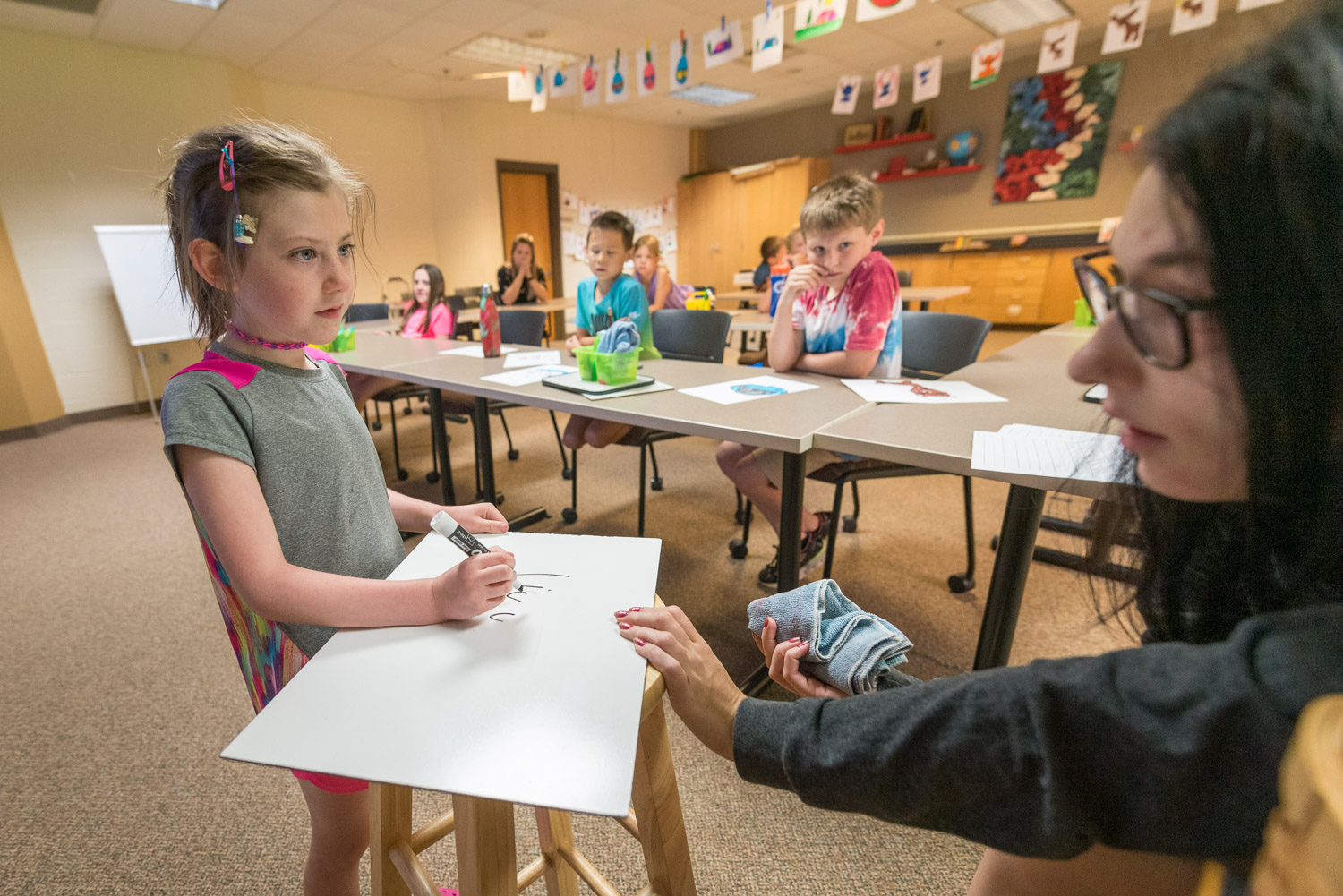 Student teacher helping student write on white board set on stool in front of classroom