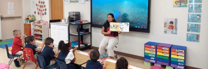 teacher in front of classroom holding book for students to see